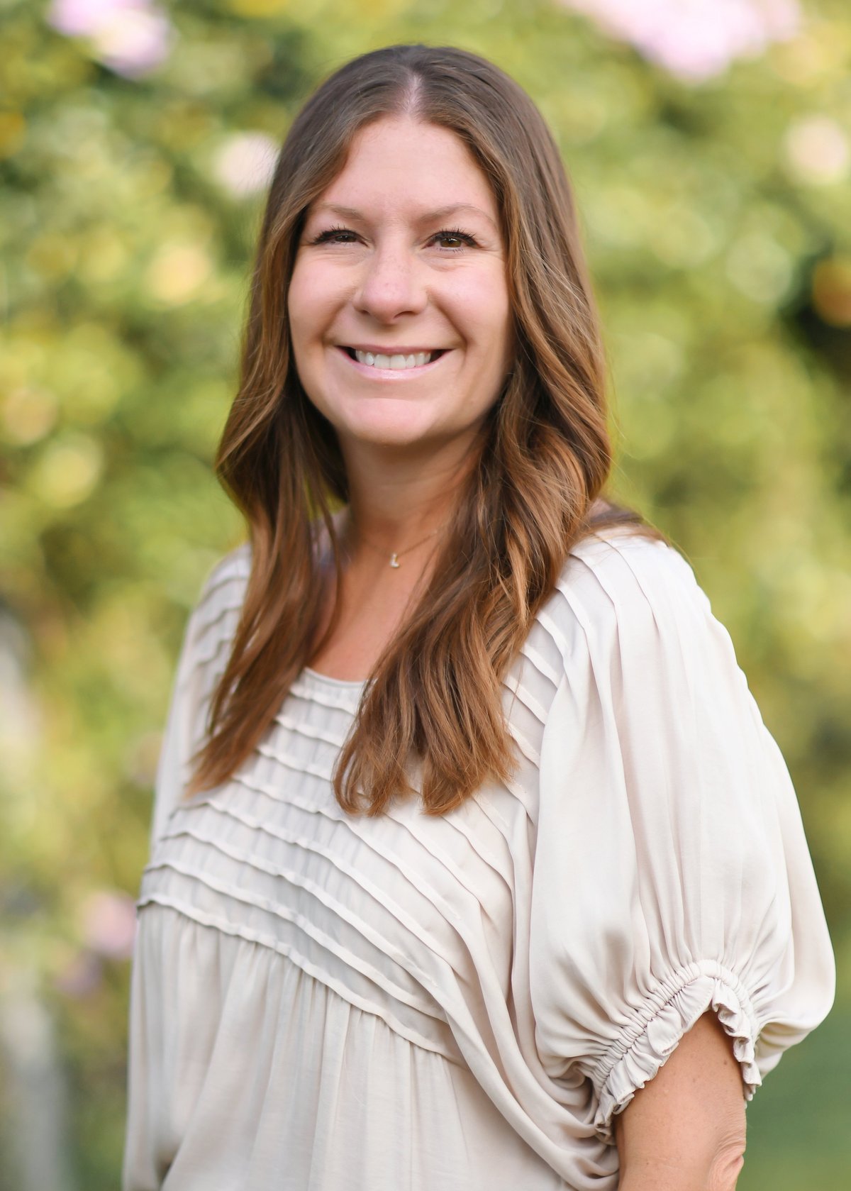Portrait of a smiling woman with long brown hair wearing a beige blouse, photographed outdoors with a blurred green background